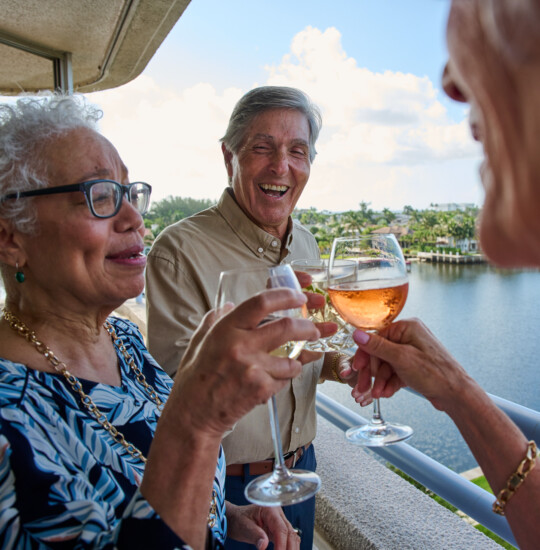 three senior friends laugh and toast wine glasses on the balcony overlooking Harbour's Edge Senior Living Community and its namesake harbor