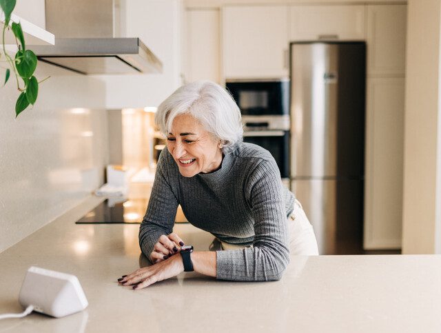 Senior woman smiling happily while using smart devices in her kitchen. Cheerful elderly woman using a home assistant to perform tasks at home.