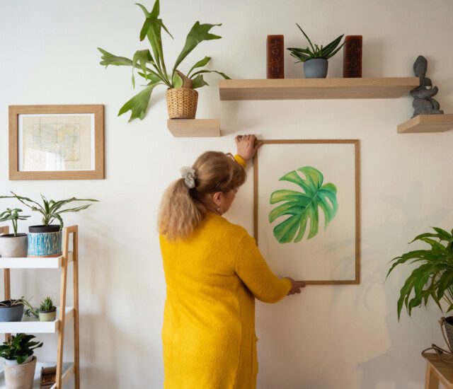 An older adult woman adjusts a colorful nature-inspired print on a wall