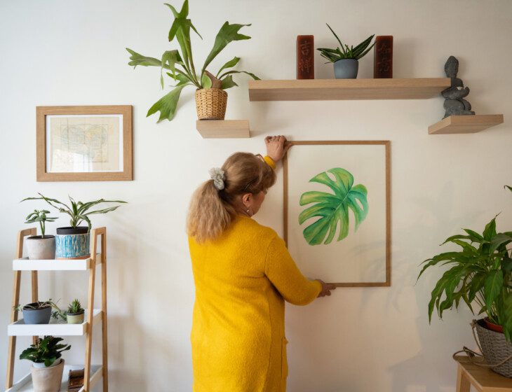 An older adult woman adjusts a colorful nature-inspired print on a wall